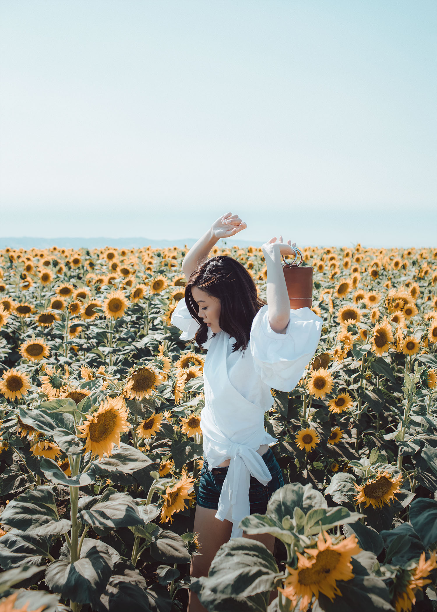 Street style fashion blogger influencer Jenny Tsang of Tsangtastic wearing white gathered ruffle puffed sleeve top denim shorts o-ring small leather bag simon miller bag in sunflower field in Davis California